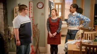 Leon Harrop as Ralph in a blue t-shirt and jeans stands in the archway of a kitchen with Sarah Gordy in a brown pinafore dress as Katie and Jamie Marie Leary in a blue and white jumper and jeans as Emma in Ralph & Katie.