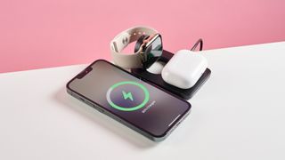 The Mous MagSafe 3 in 1 charger is pictured open on a white tabletop with a pink background. An iPhone, Apple Watch, and Air Pod case are shown charging.