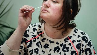 white woman wearing white sweater with colorful animal print tilts her head back in order to insert a long swab into her nose.