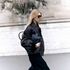 Model wearing sunglasses and a black collared jacket holding a black tote handbag under her arms sold at Luisaviaroma