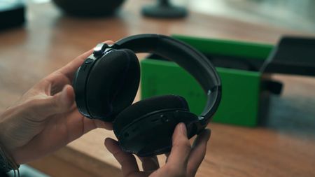 A person holds a pair of black Razer Project Motoko wireless headphones, showcasing their design, with a green box visible in the background on a wooden surface.