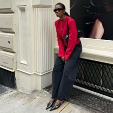 A woman wears a red jacket, black jeans, black slingbacks, and the alaia le teckel bag in black.