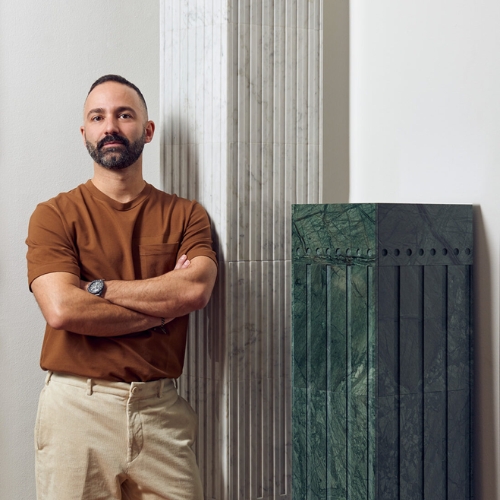 Image of a man in a burnt orange t-shirt and khaki pants standing with his arms folding leaning against a marble pillar.