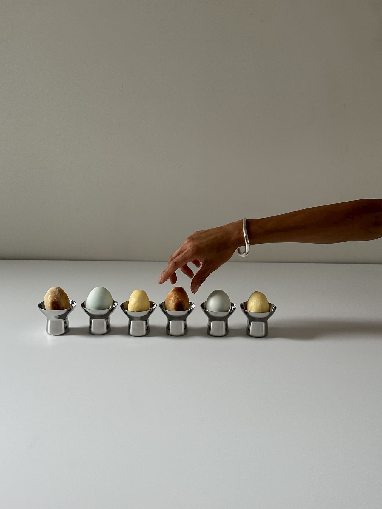 A hand with a silver bracelet on it is captured fixing a series of eggs sitting in silver eggcups.