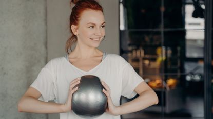 woman wearing a white tshirt holding a small grey pilates ball in grey stone wall background setting. 