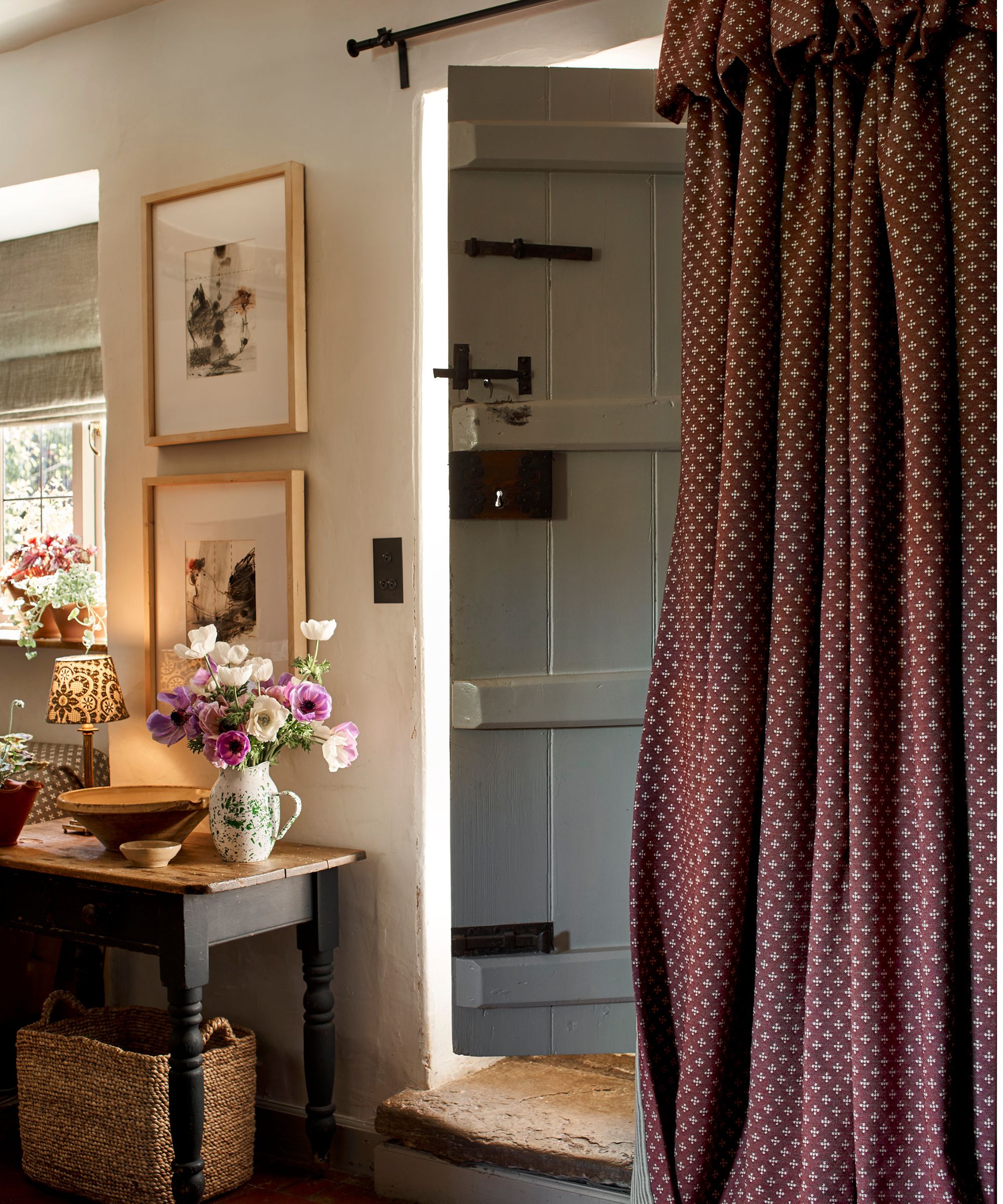 cottage entryway with a barn door with a red flop over frill curtain, antique small table styled with artwork, florals in a vase and a small lit lamp
