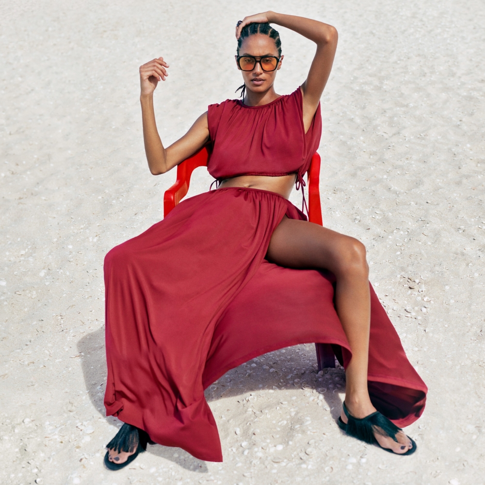 Model wearing a deep red two piece top and skirt set sold at H&amp;M with black fringe sandals and yellow lens sunglasses on a red plastic chair on the beach