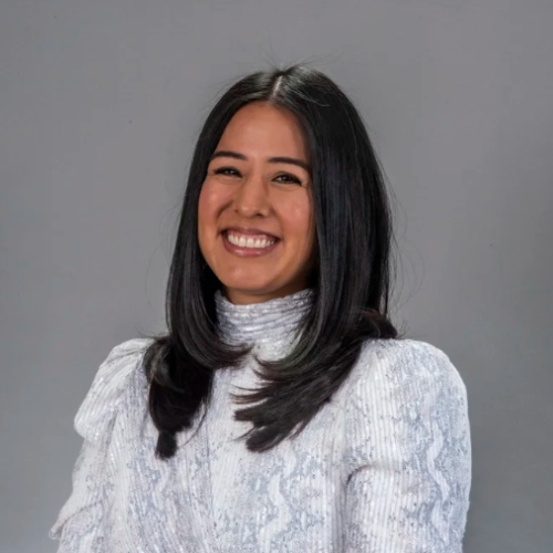 Image of a woman in a white and gray blouse sitting in front of a gray background.