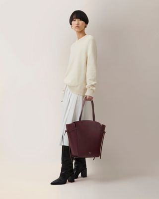 Woman wears cream jumper, white skirt, black boots and burgundy bag