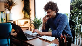 Person on their phone at a laptop at a desk, working