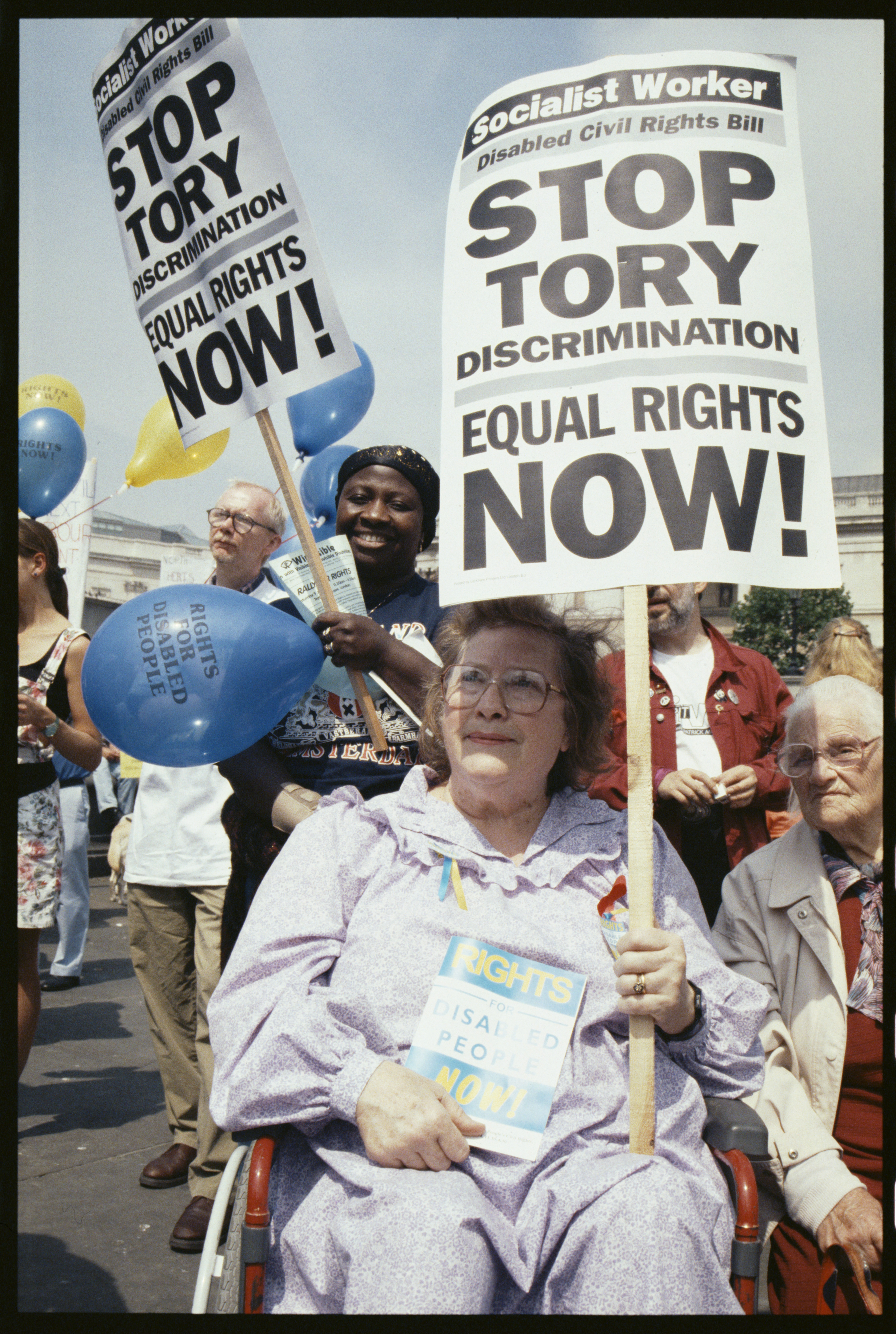 A woman in a wheelchair holds a placard reading "Stop Tory discrimination, equal rights now!" at a sunny Trafalgar Square rally, with balloons reading "Rights for disabled people" visible behind her.