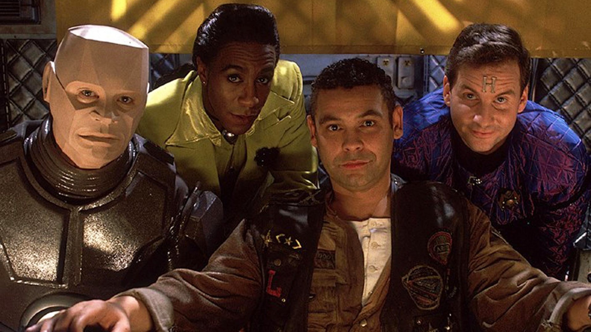 Still from the British sci-fi sitcom called Red Dwarf. Here we see four of the main cast sitting down. From left to right: Kryten, the Cat, Lister, and Rimmer.