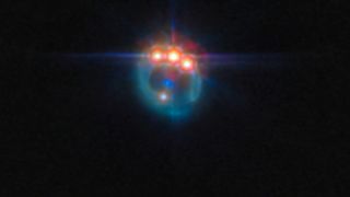 A small image of a galaxy distorted by gravitational lensing into a dim ring. At the top of the ring are three very bright spots with diffraction spikes coming off them, right next to each other: these are copies of a single quasar in the lensed galaxy, duplicated by the gravitational lens. In the centre of the ring, the elliptical galaxy doing the lensing appears as a small blue dot. The background is black and empty