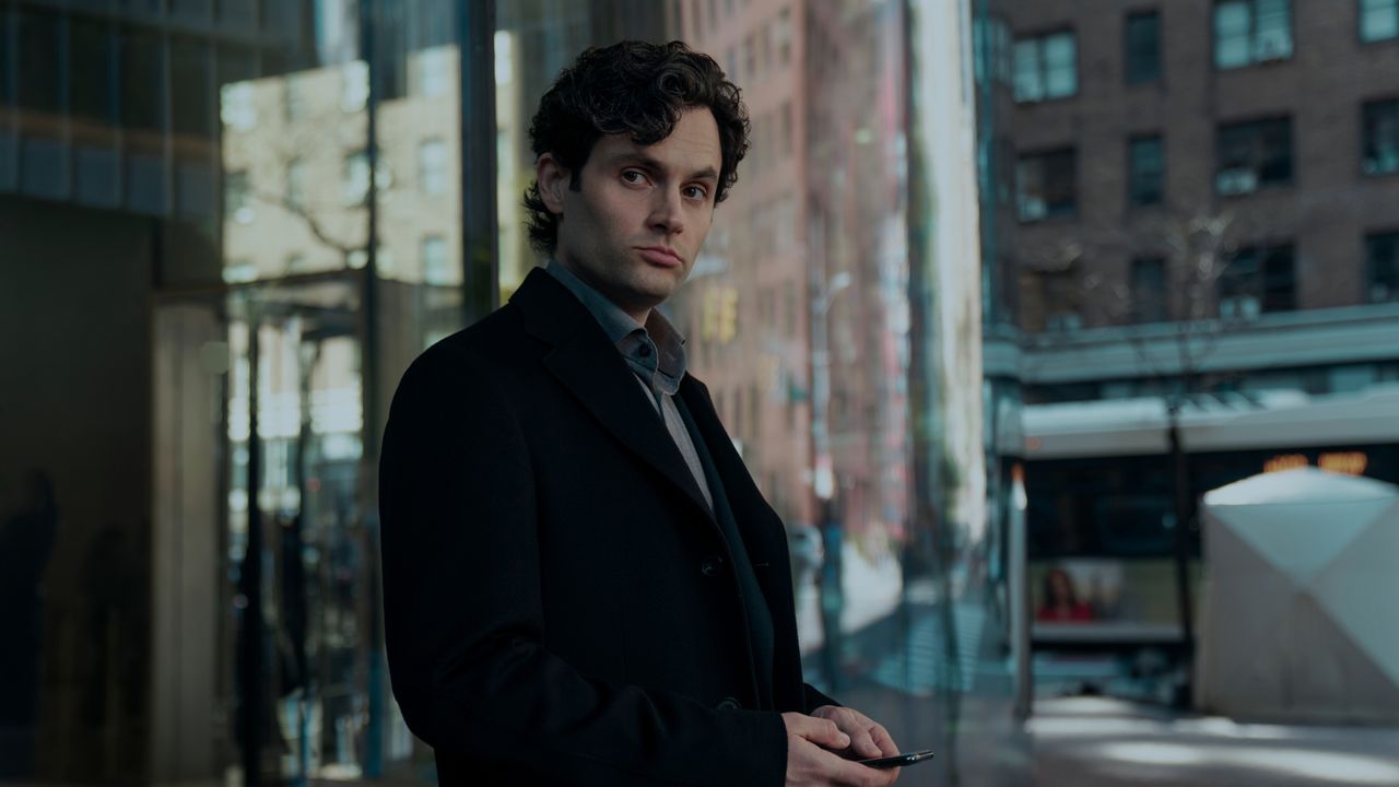 Penn Badgley as Joe Goldberg, standing outside an NYC high-rise building while holding a cell phone, in episode 502 of You.