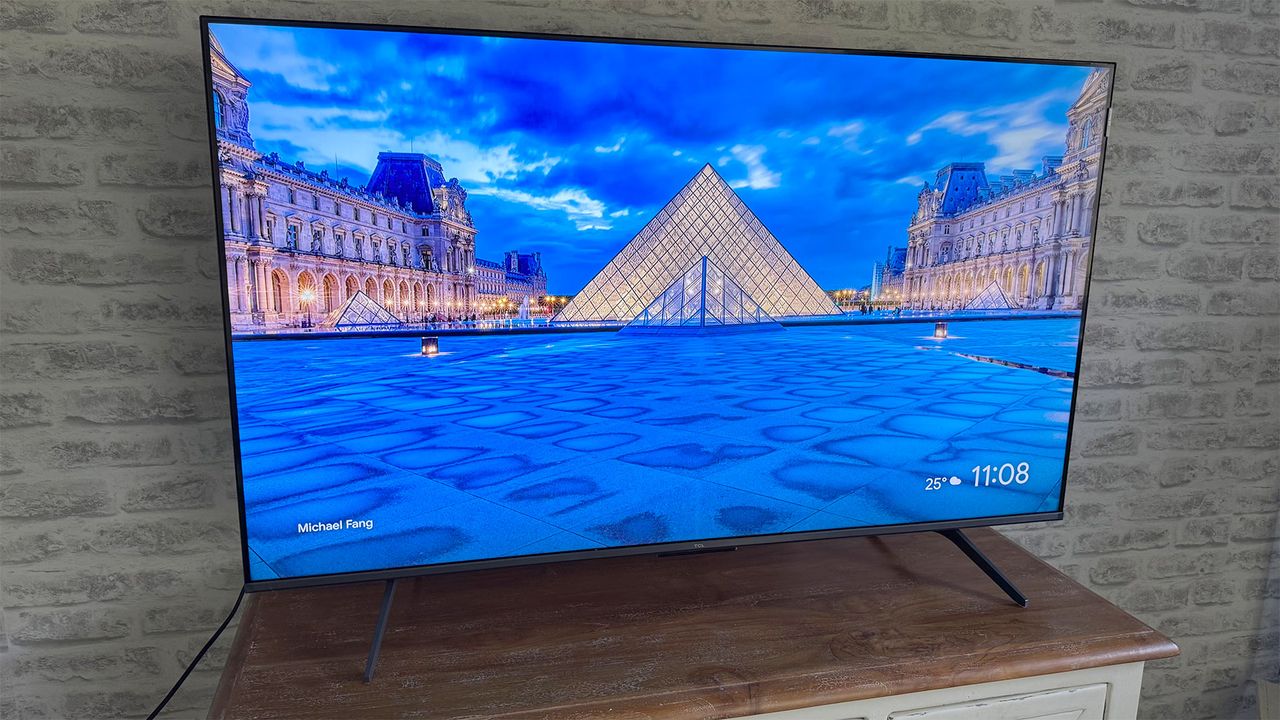The TCL C6KS 50-inch TV on a TV cabinet, seen at a slight angle, in front of a brick wall. On screen is the exterior of the Louvre museum in Paris at night.
