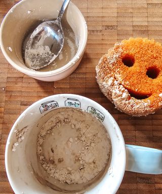 A birds eye view of a mug with baking soda in it, an orange Scrub Daddy, and a ceramic pot with a spoon in it, all on a wooden chopping board