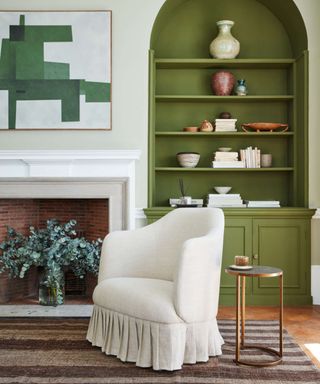White skirted armchair in front of fireplace and green arched alcoves with brown striped rug