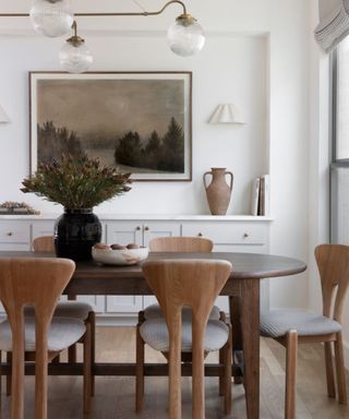 a white dining room with an oval wooden dining table, shapely wooden dining chairs and brown accessories including artwork and a decorative jug