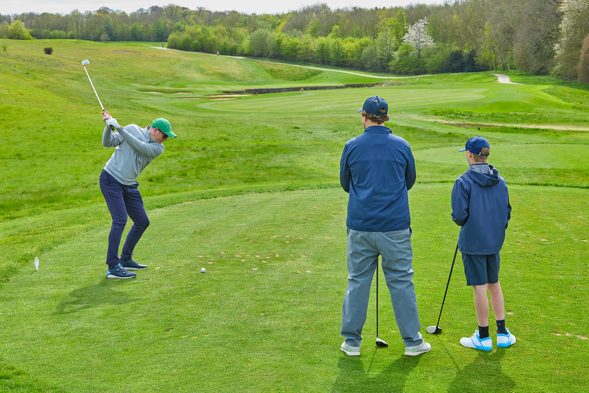 A golfer hitting a long iron off the tee with two playing partners watching on
