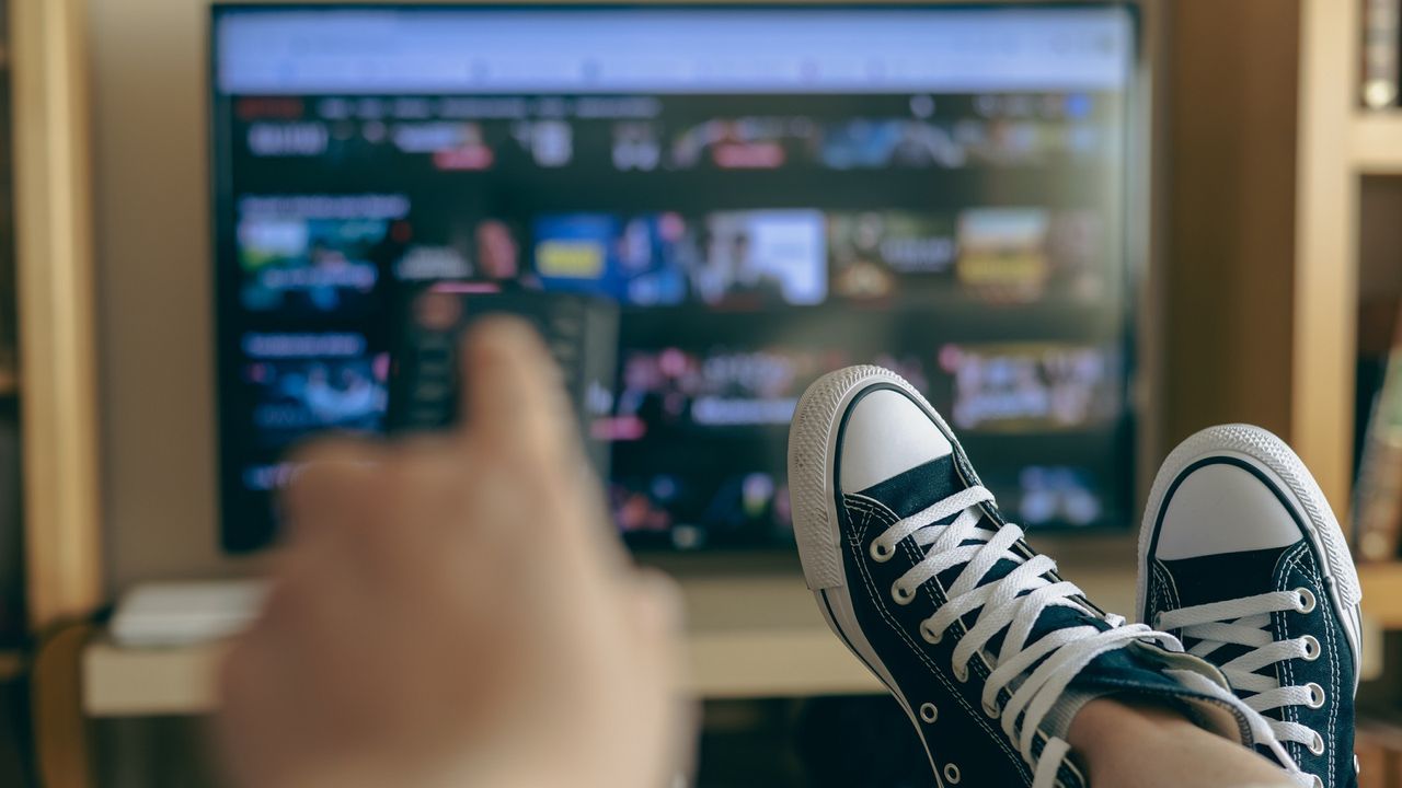 Someone&#039;s hand holding a remote, with sneakered feet up and streaming TV in background.