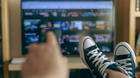 Someone's hand holding a remote, with sneakered feet up and streaming TV in background.