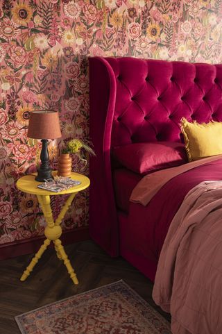 A bedroom with pink wallpaper, a velvet studded pink headboard and a yellow side table
