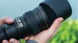 Nikon AF-S 300mm f/4E PF ED VR held in the hand while attaching lens hood in front of green and blue outdoor setting