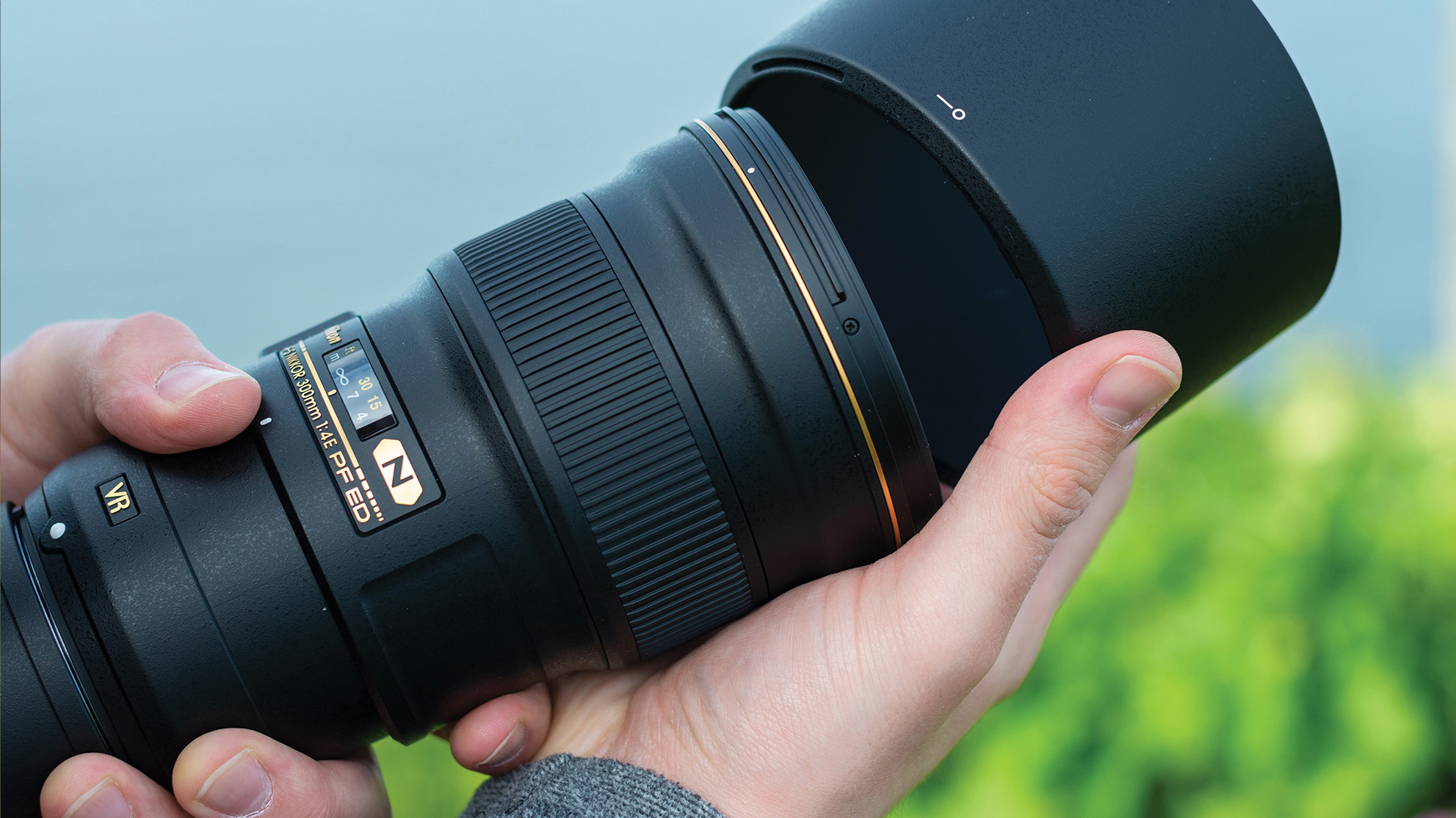 Nikon AF-S 300mm f/4E PF ED VR held in the hand while attaching lens hood in front of green and blue outdoor setting 