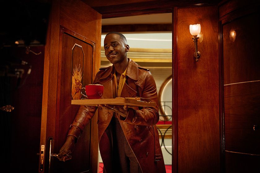 The Doctor is walking through an ornate mahogany door into a hotel room, carrying a wooden lunch tray, on which is a red mug with foam visible on the top, and a plate with a toasted sandwich.