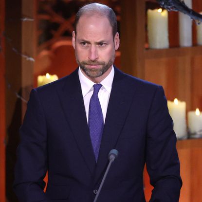 Prince William wearing a blue suit and purple tie standing at a mic in front of lit candles