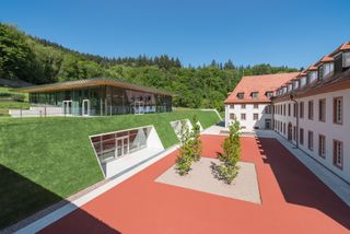 United World College by Peter Kulka Architektur and Hotz + Architekten. A large three storey building next to a courtyard made of red bricks next to a building built into a green hill.