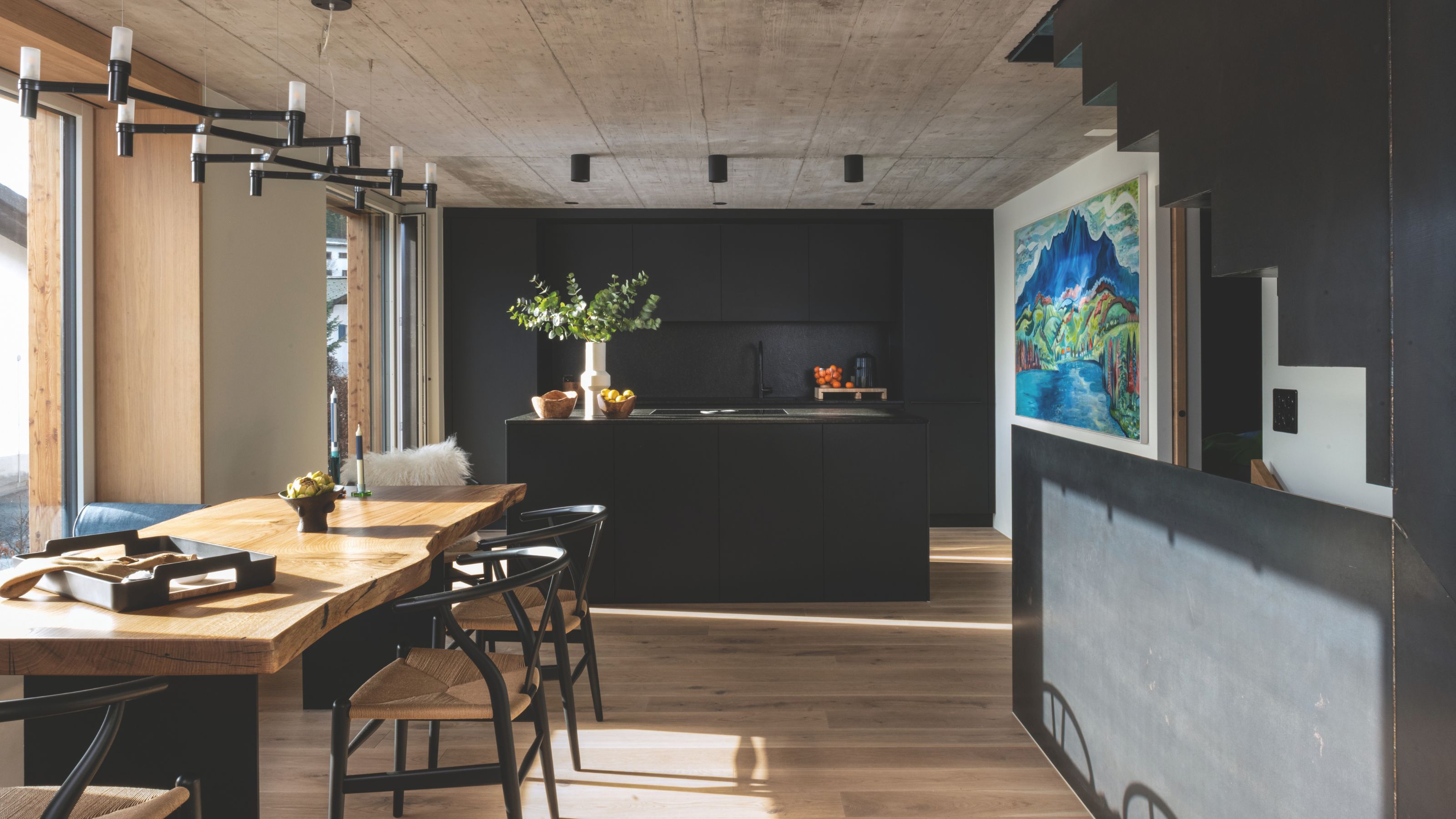 A modern open-concept kitchen and dining area featuring a live-edge wooden table, black wishbone chairs, and a sleek matte black kitchen island under a concrete ceiling