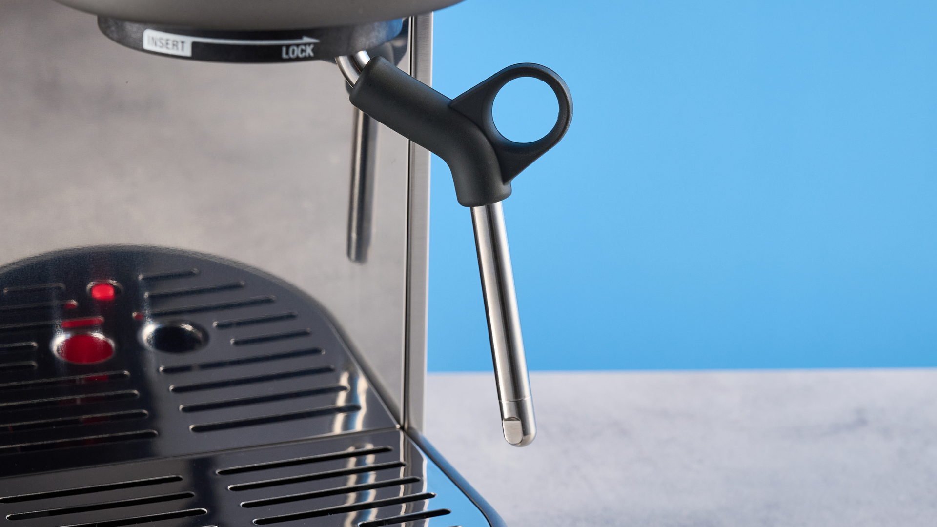 the breville bambino / sage bambino in silver photographed against a blue background with a wooden feature wall, showing the steam wand, group head, water tank, button controls, and espresso