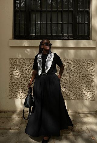 A woman wearing a black t-shirt with a black and white scarf, black belt, a voluminous black skirt, and black ballet flats.