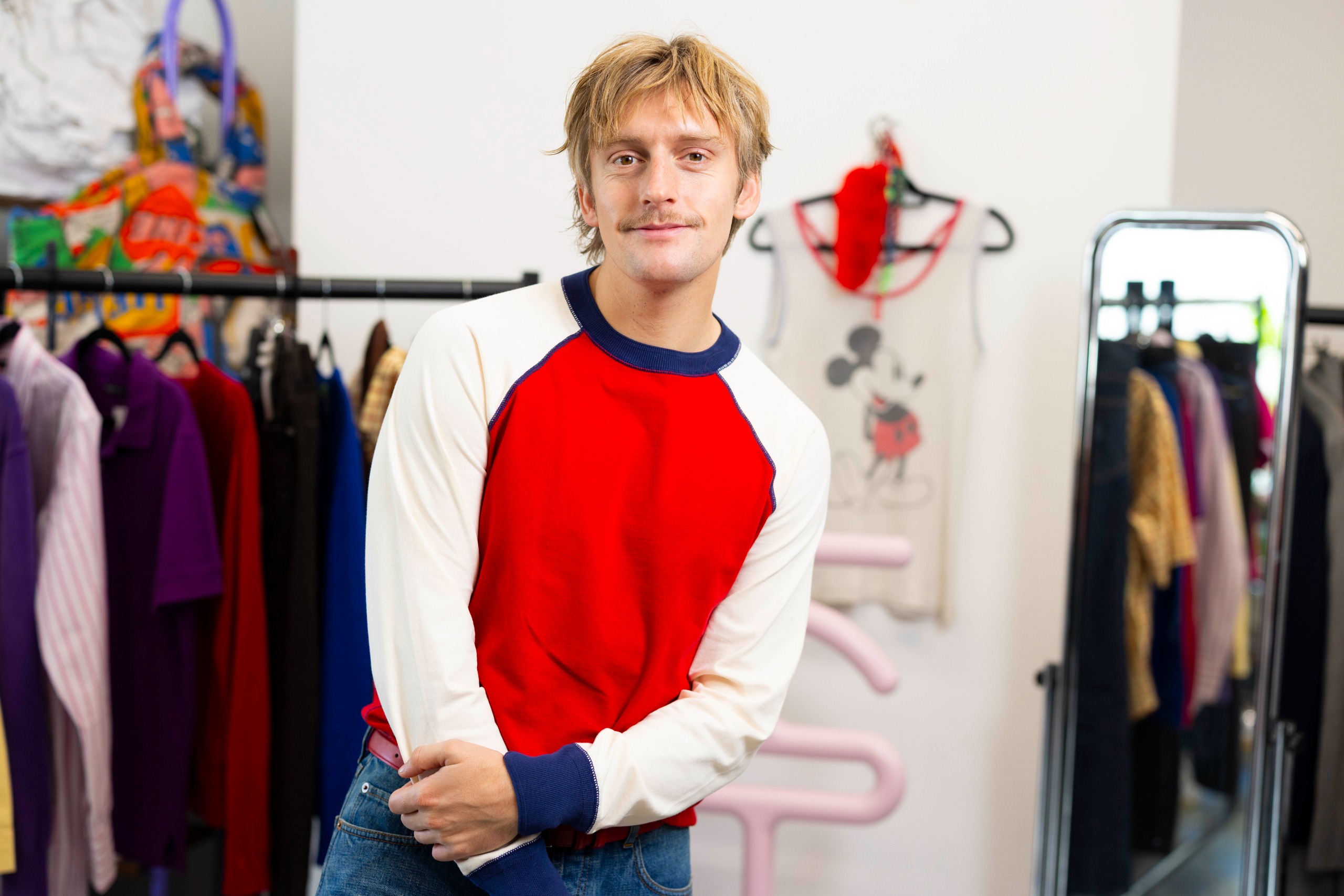 A young blonde man in a red, blue, and white long-sleeved jumper, standing in a filled-with-colorful-clothing studio.
