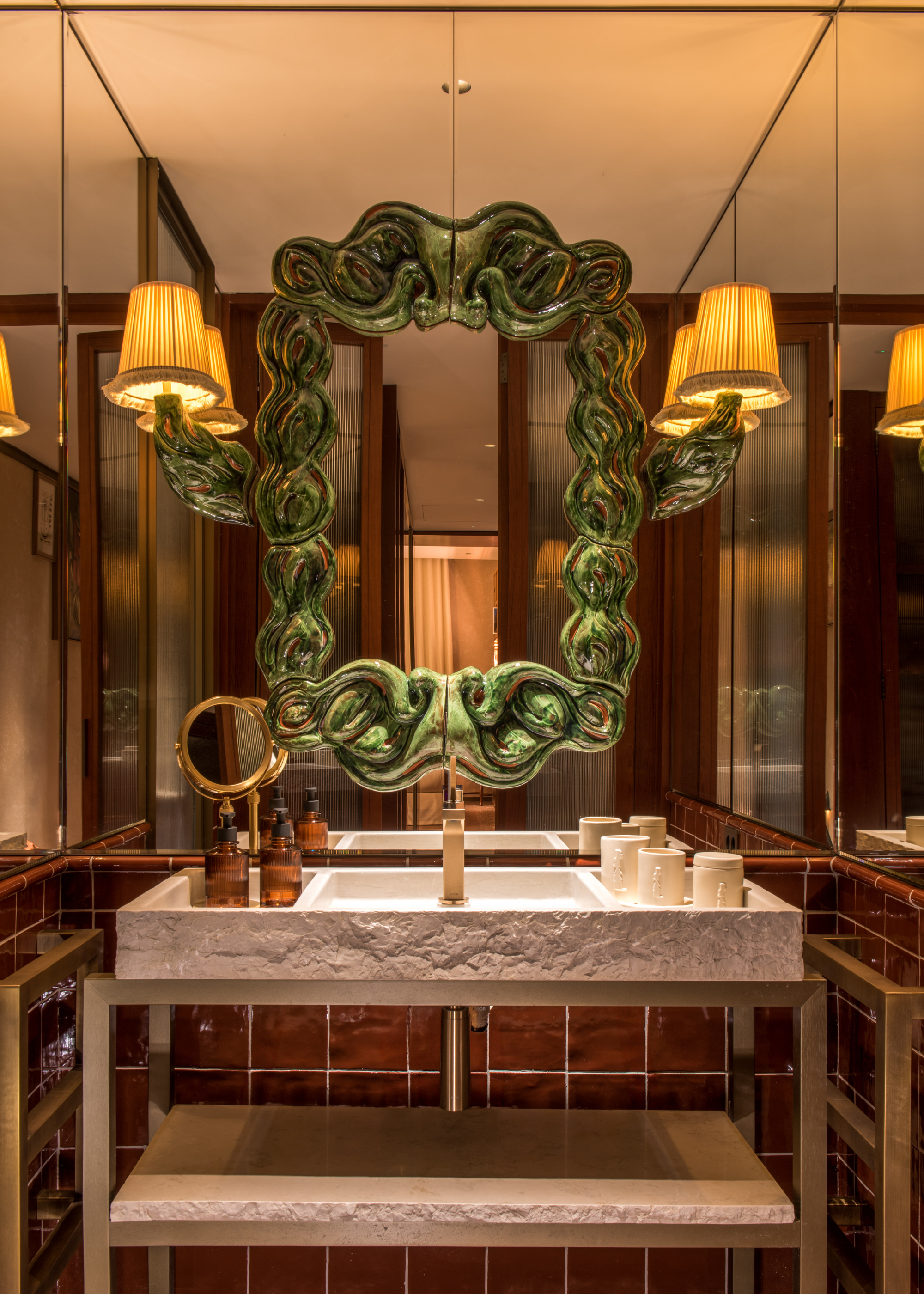 A statement sculpted green mirror with a set of attached lamps over a wash basin in a tiled bathroom