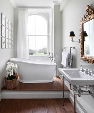 An ensuite with a white free-standing bath in front of a window on a raised section of wooden flooring