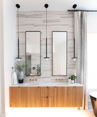 A double bathroom vanity with tall slim black mirrors, three hanging pendant lights, two sinks with gold faucets, a wooden base, and warm colored lights glowing underneath the sink