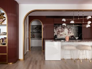 Cream marble counters with red veining, with flutted walls in deep burgundy and cream, tied together with gold detailing and a white kitchen islan