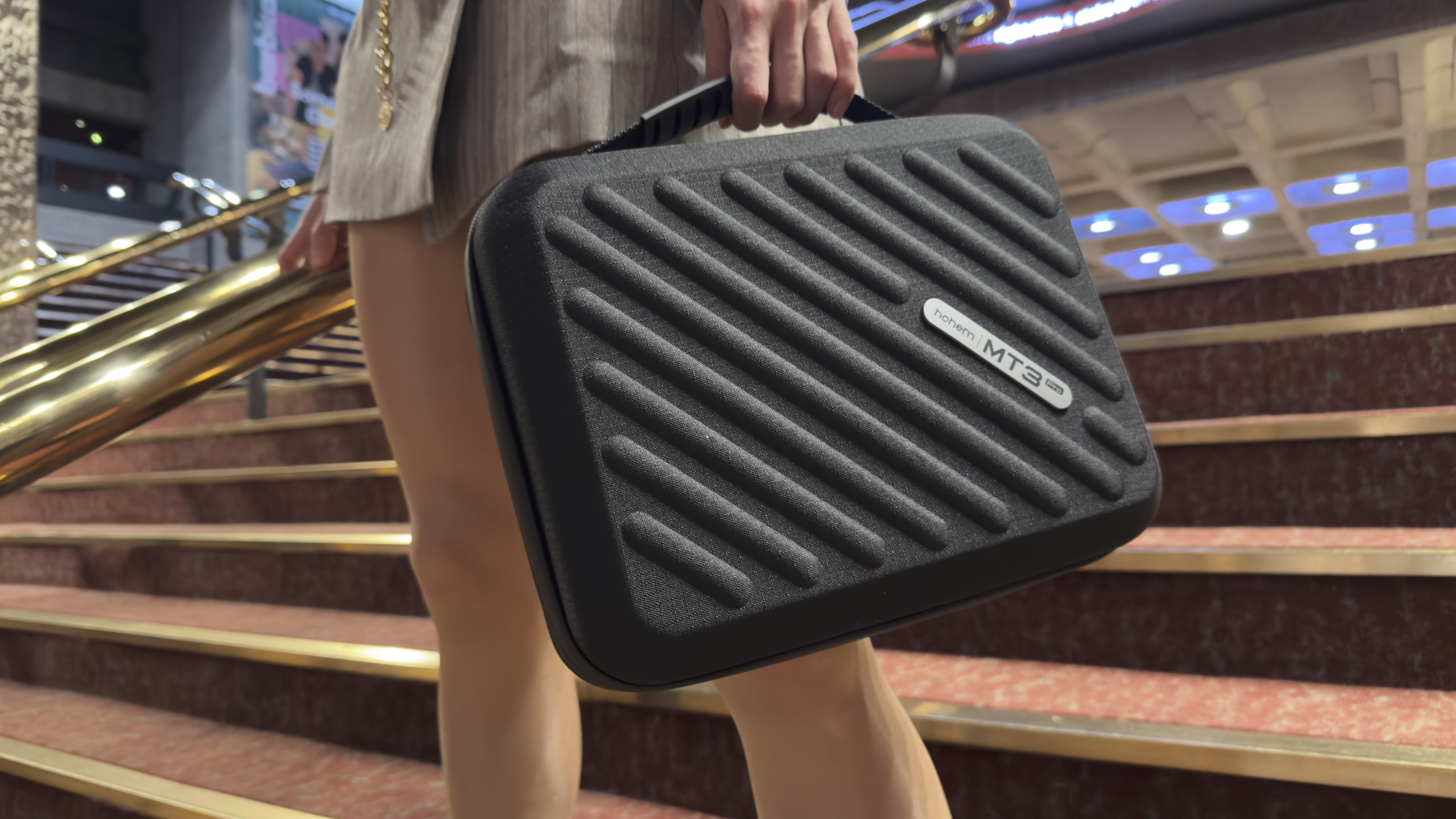Person carrying a black Hohem MT3 hard carry case with textured ridges while walking up indoor stairs.