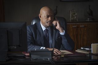 Vic Wallinger (Laurence Fishburne) sits at a desk with a computer in front of him, making a call on a landline phone. He is wearing a blue suit with a lighter blue shirt and a paisley tie, and looks concerned