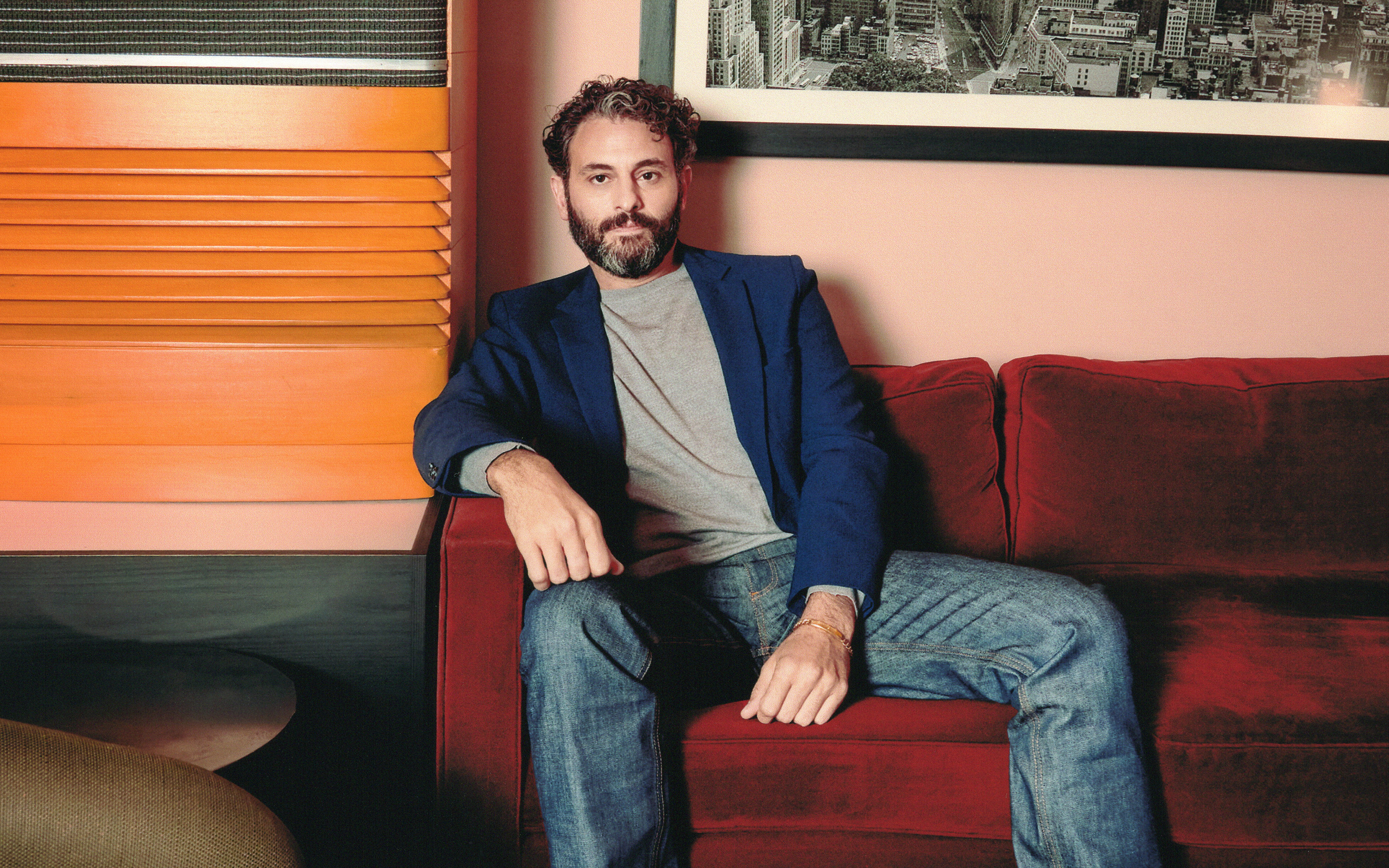 Arian Moayed wears jeans and a blue blazer leaning back sitting in a red velvet couch