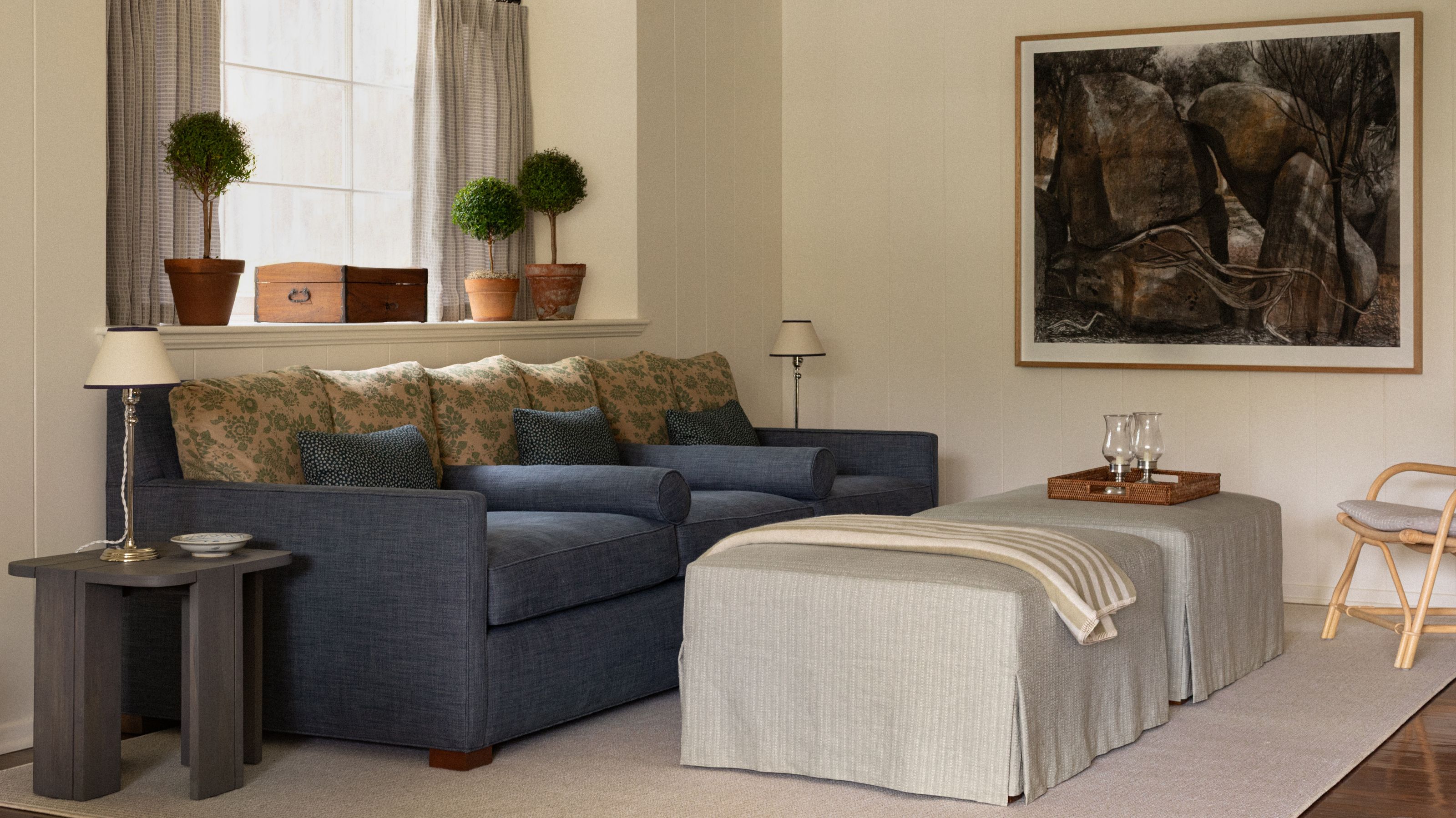 a warm neutral living room corner with a large window, a navy couch with yellow floral back pillows and two striped ottomans