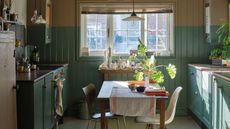Small kitchen with sage green cabinets on left and right, with window at middle back, and table in middle fo space with two mismatched chairs.