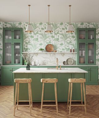 A kitchen with green and white leafy wallpaper, green cabinets, a kitchen island with white marble surfaces and a green base, three light wooden chairs and three gray pendant lights above it