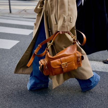 Person in wide-leg jeans, a trench coat, and boots walking in the street holding a leather handbag