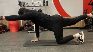 Man demonstrates bird dog exercise. He balances on his right hand and left knee, with his left arm and right leg extended and raised off the floor. He wears Nike trainers, black pants, black sweatshirt and black cap.