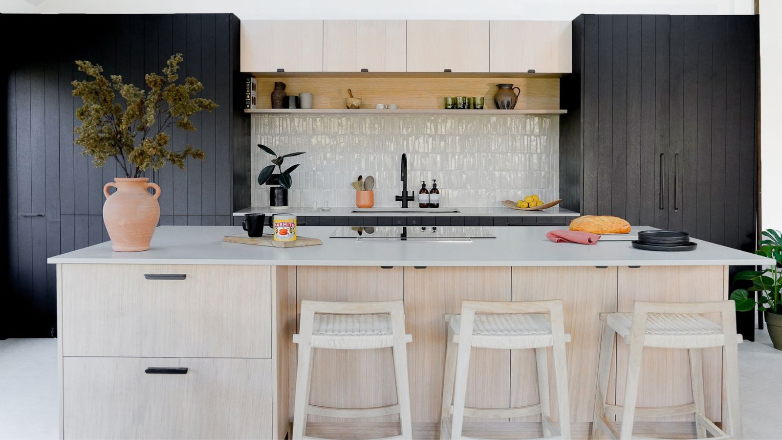 A pale wood kitchen island with a sleek worktop creates a bright focal point, paired with light stools for relaxed seating. Dark cabinetry and a textured tiled splashback add contrast and depth.