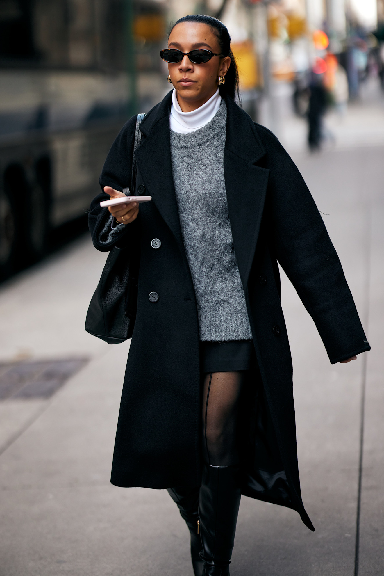Lauren Tappan wearing a black coat, grey crewneck sweater, white turtleneck sweater, black skirt, tights, and black boots