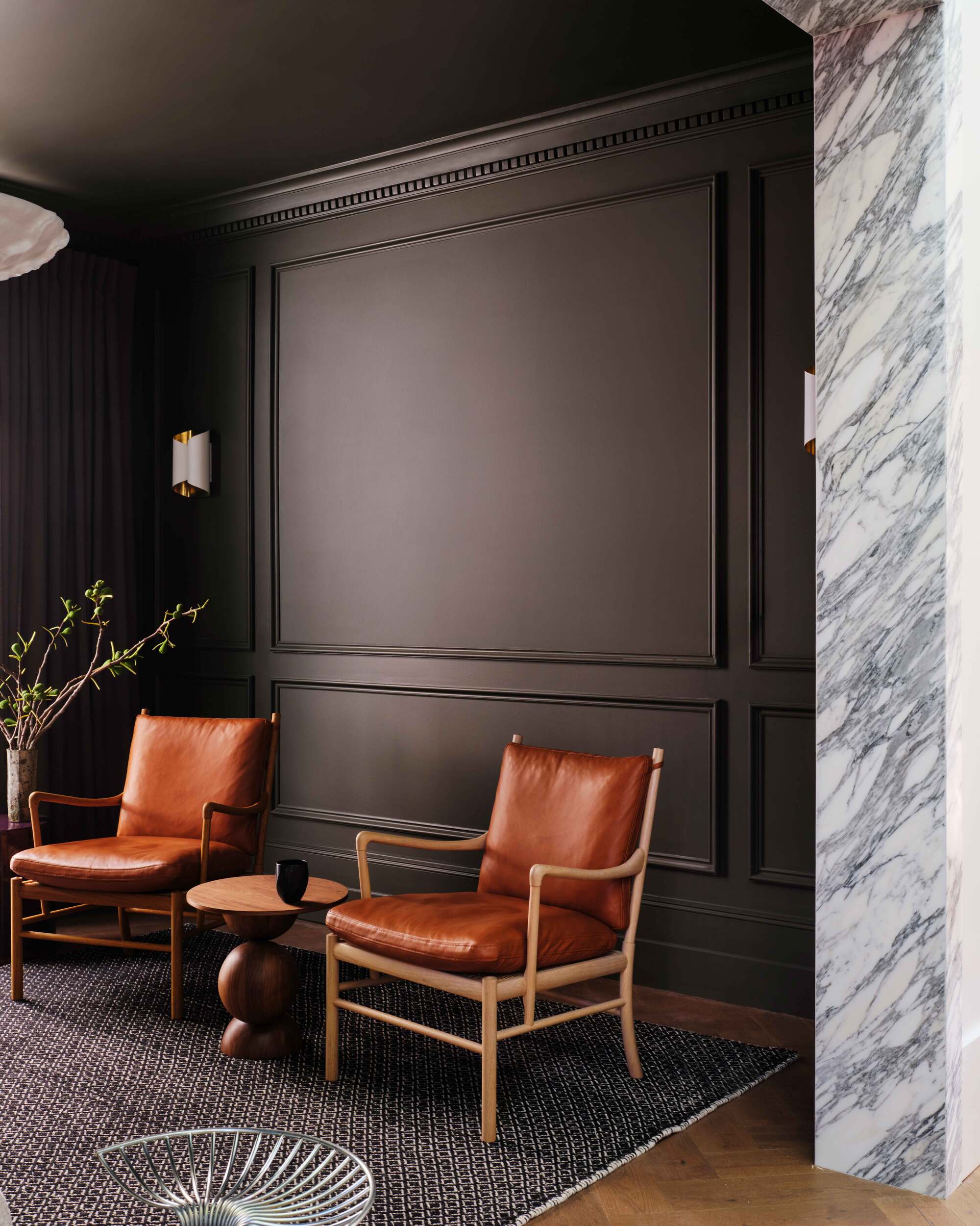 Image of the corner of a dark gray/black living room with crown molding on the walls. There are two orange, leather accent chairs with a small wooden side table between them. There is a dark gray rug on the floor and a green plant in the corner.